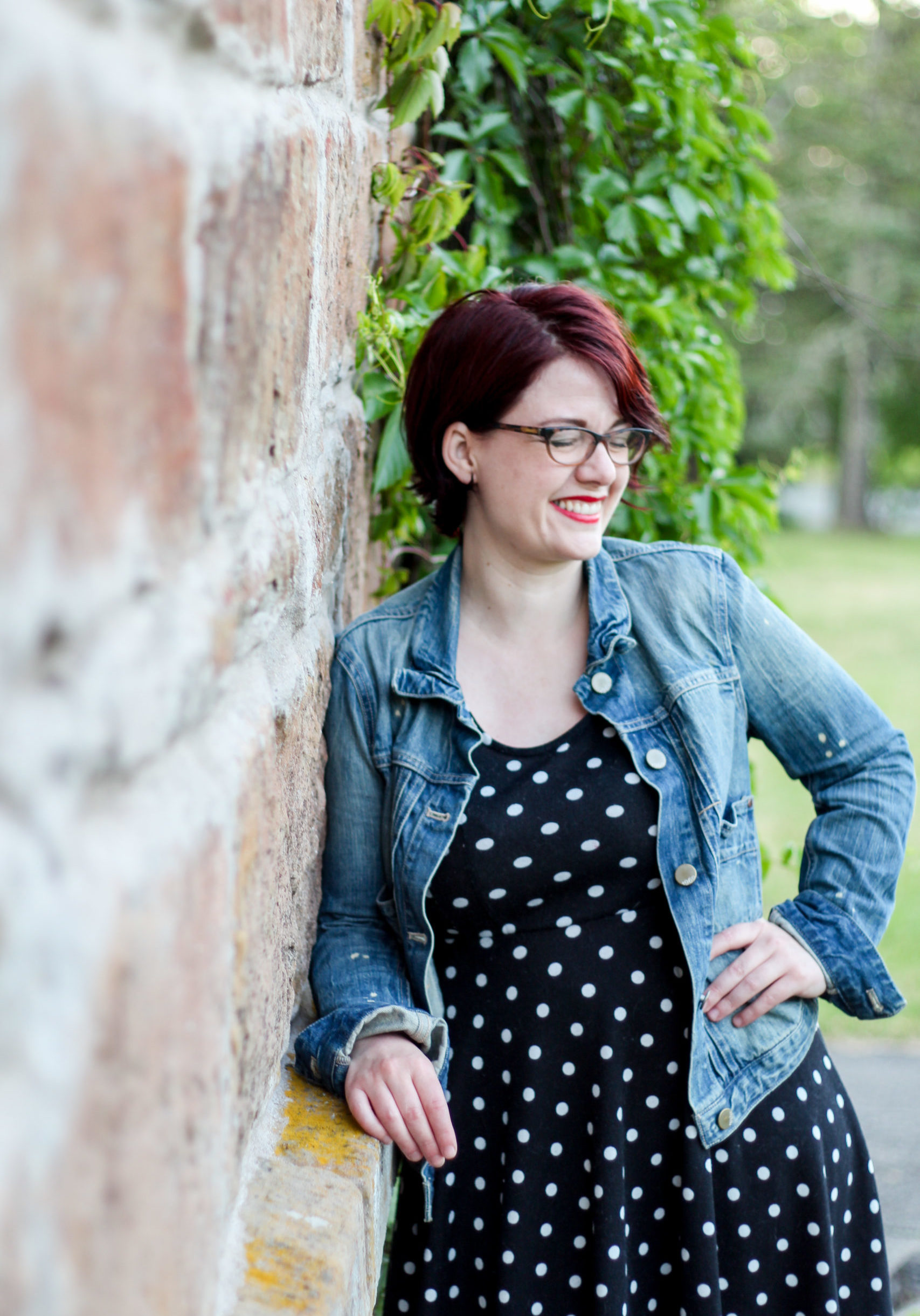 Kris is leaning against an old brick wall, laughing. She is wearing a worn jean jacket and black and white polka dot dress, with short auburn hair and glasses.